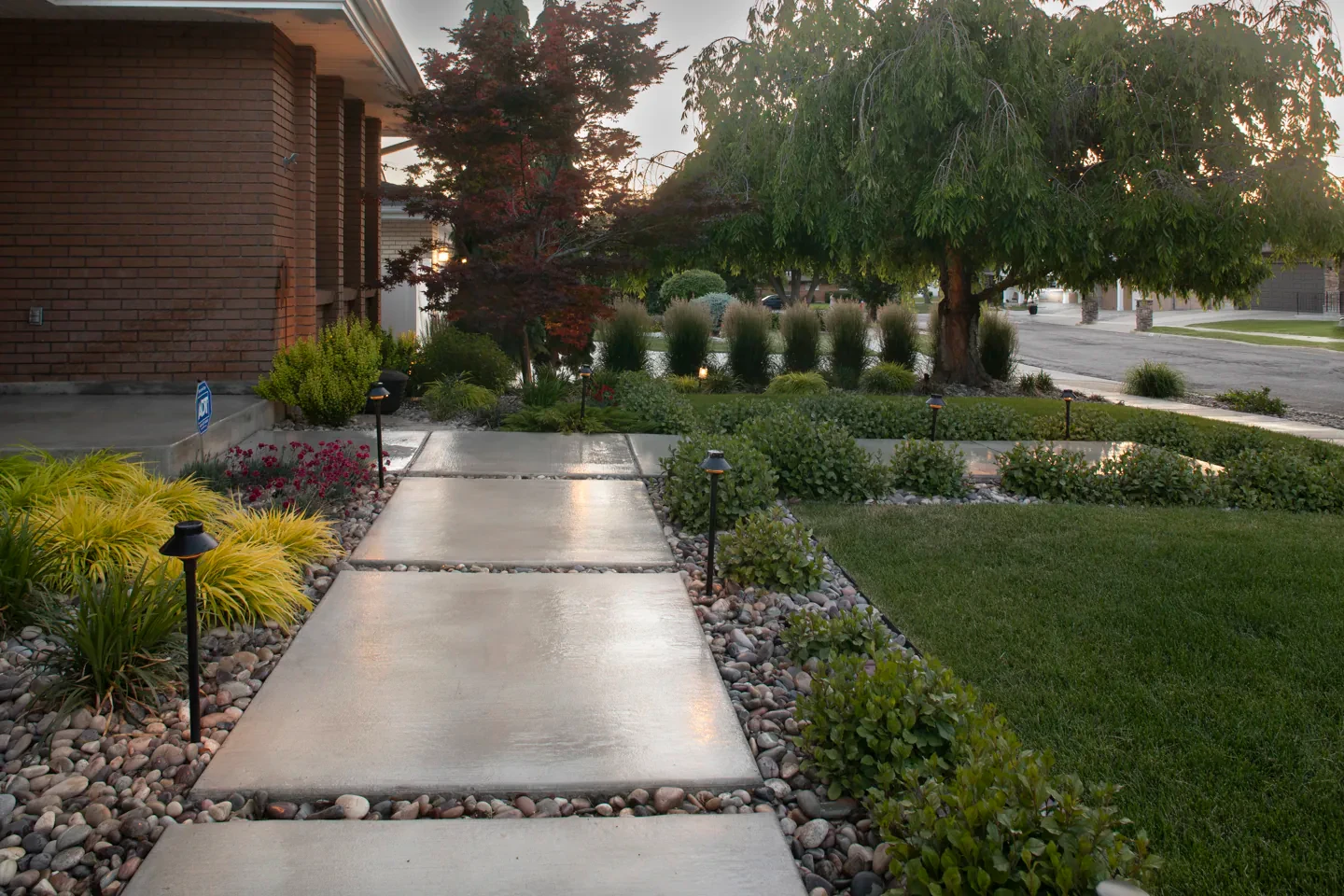Low-voltage path lights illuminate a concrete front walkway, planting beds and lawn in Millcreek at dusk