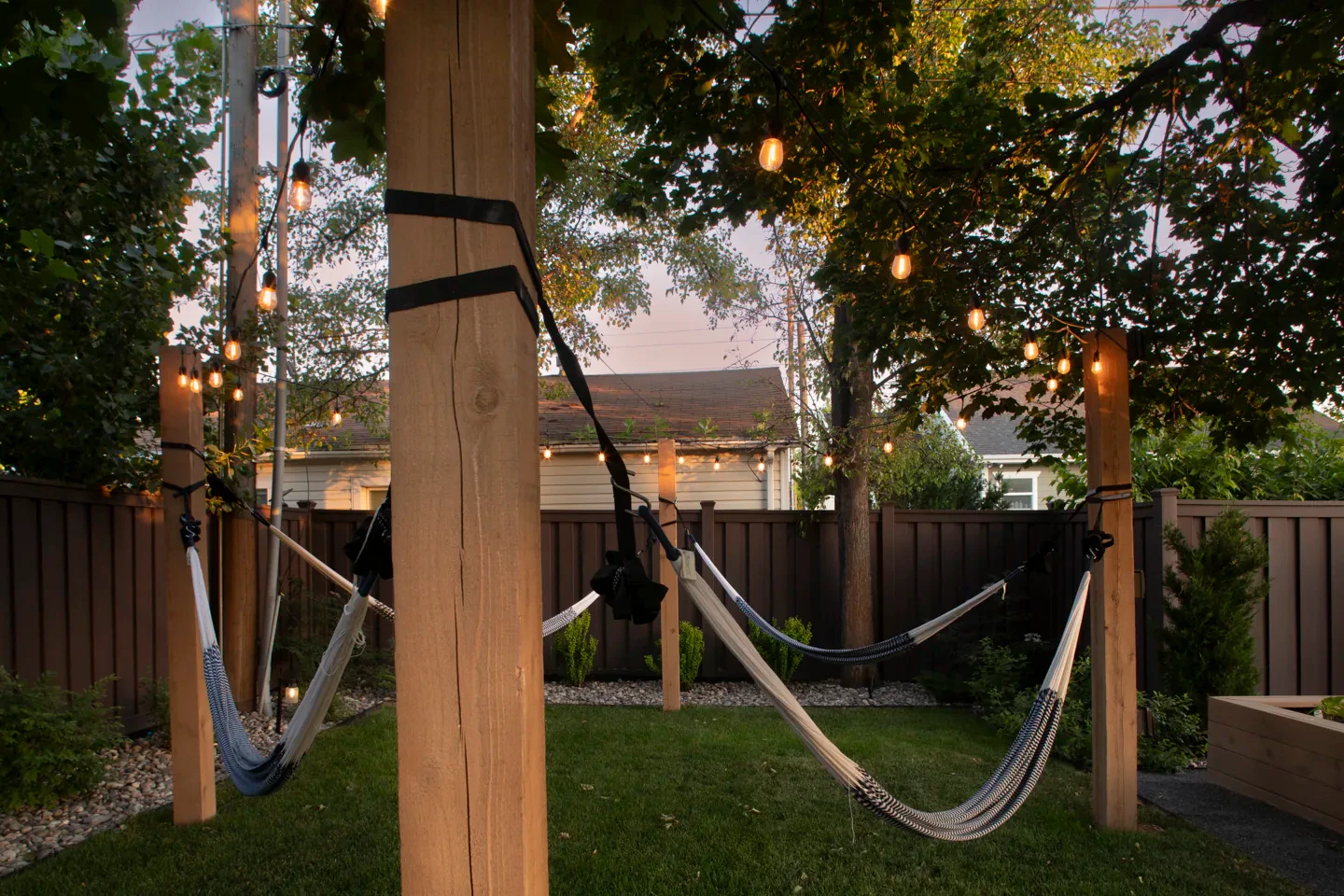 Twin hammocks hung beneath a Millcreek pergola with bistro lights and a private backyard lawn