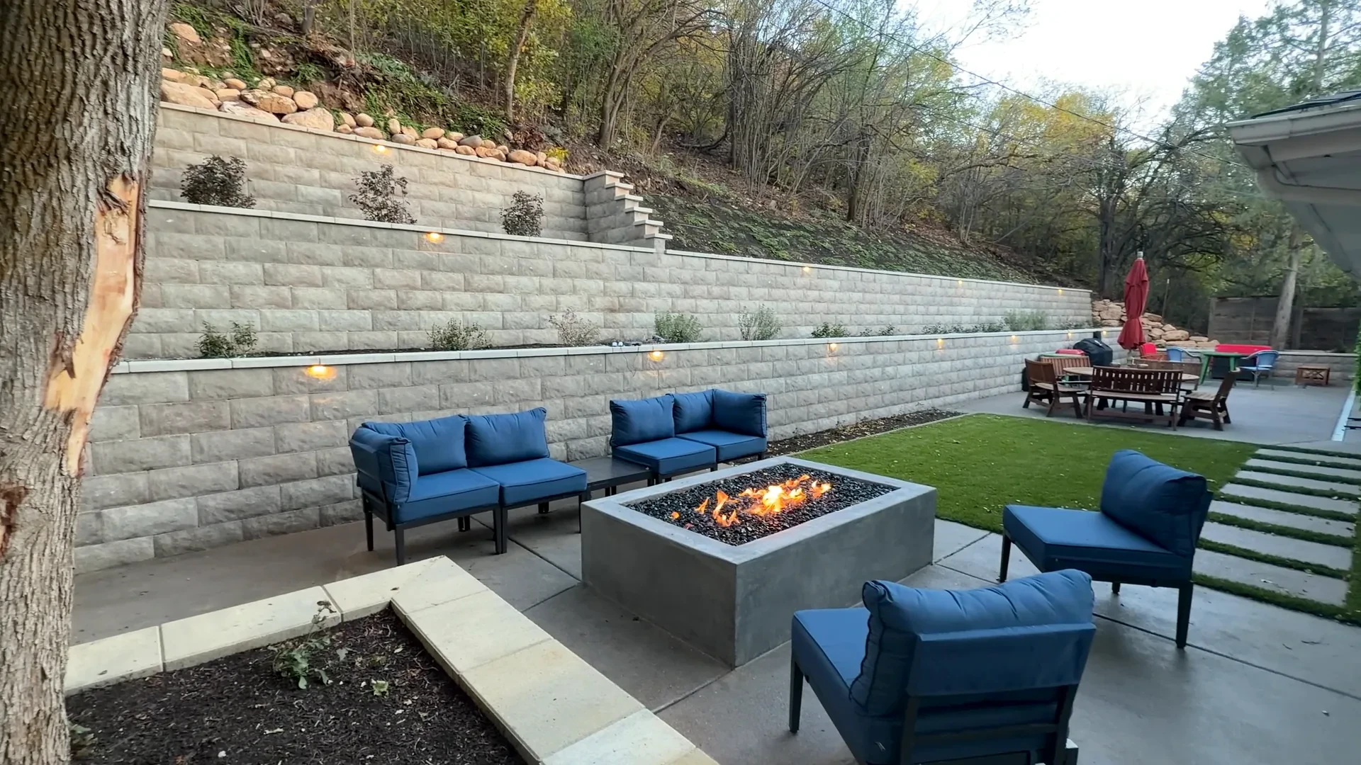 Completed Olympus Cove backyard at dusk — terraced Belgard walls, pewter exposed-aggregate patio and polished concrete gas fire feature