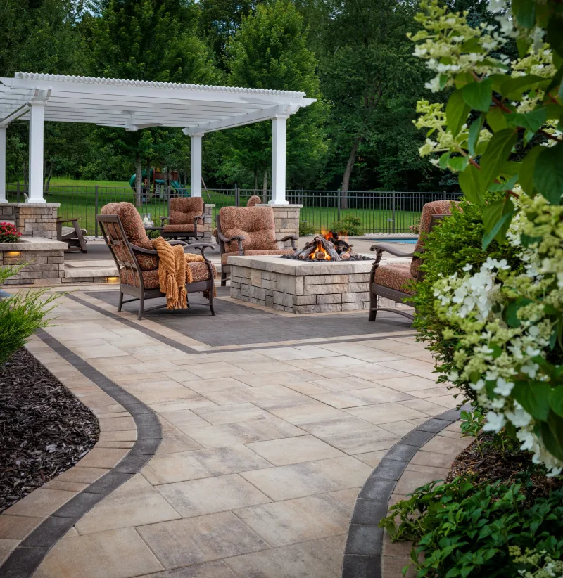 Belgard Dimensions paver patio with curved dark-border inlay, Weston Stone fire pit surround, Ashlar Tandem Wall arbour gate, and lounge seating — manufacturer inspiration.