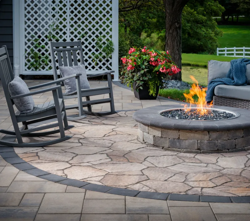 Belgard MegaArbel circular patio inset with Brooklyn Paver border, Weston Stone fire pit with glass media, rocking chairs, wicker sofa, and white lattice pergola — manufacturer inspiration.