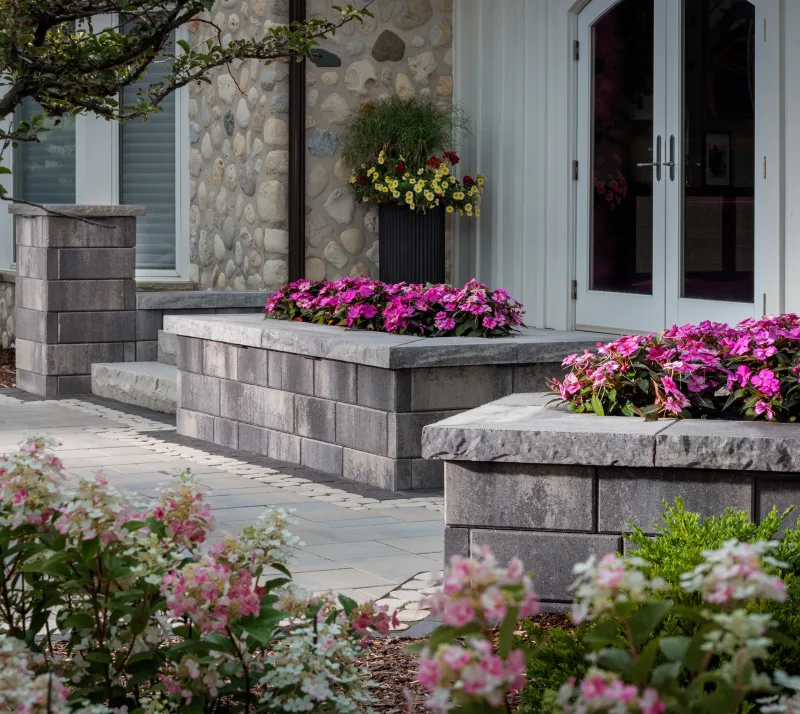 Belgard Melville Tandem Wall raised planter boxes with charcoal coping caps, Dimensions 18 walkway with cobble inlay, vivid pink impatiens, and river cobble stone veneer — manufacturer inspiration.