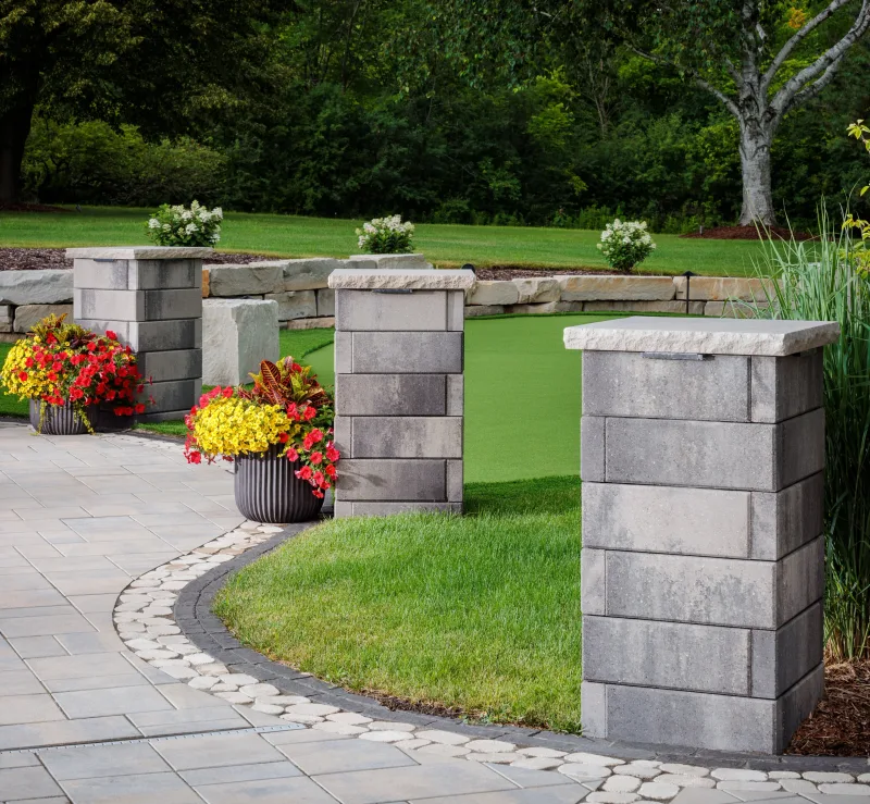 Belgard Melville Tandem Wall columns with natural stone slab caps, curved cobble border, Dimensions 18 paver field, putting green, and mature tree canopy — manufacturer inspiration.