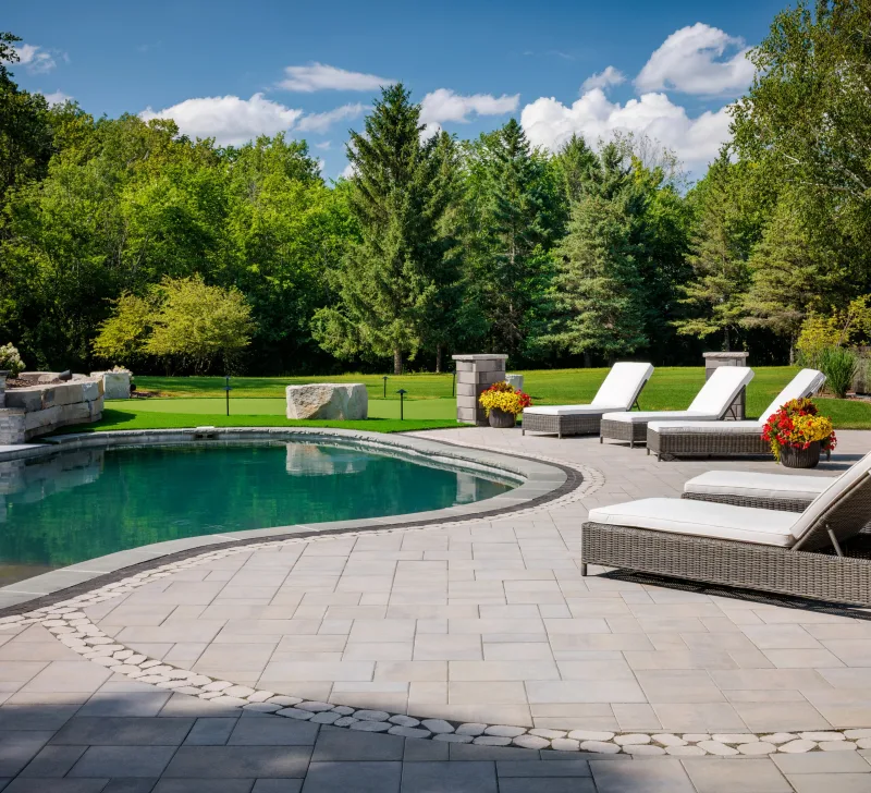 Belgard Dimensions 18 pool surround with curved cobble mosaic border, Melville Tandem Wall columns with planters, lounge chairs, and putting green — manufacturer inspiration.
