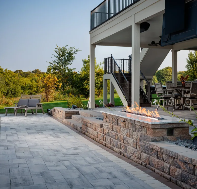 Belgard Dimensions 12 paver patio with Highland Stone fire pit surround and lake backdrop — manufacturer inspiration.
